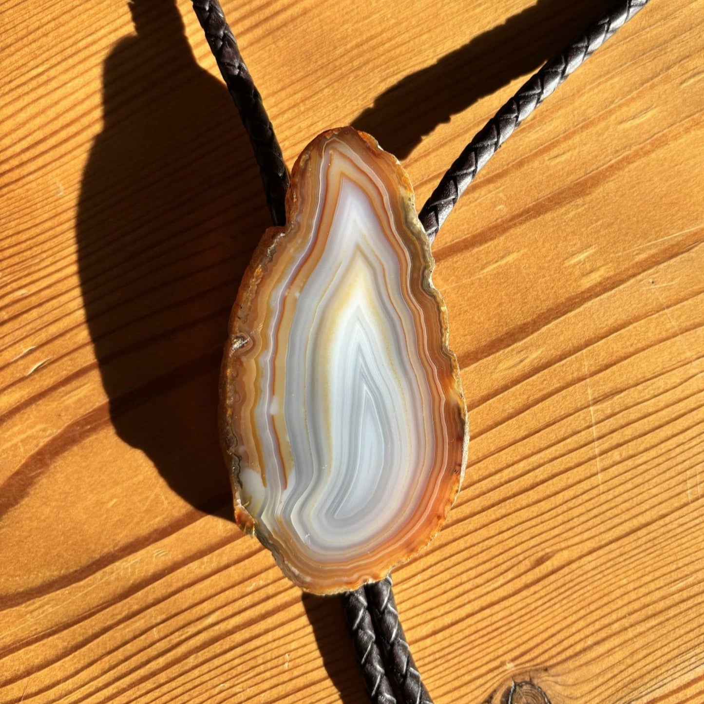 The Cimmaron Bolo Tie by My Store features a polished, colorful agate slice with brown, white, and gray bands. Its set on a wooden surface with a braided leather cord and pewter tips. The sunlight enhances its natural beauty and texture.