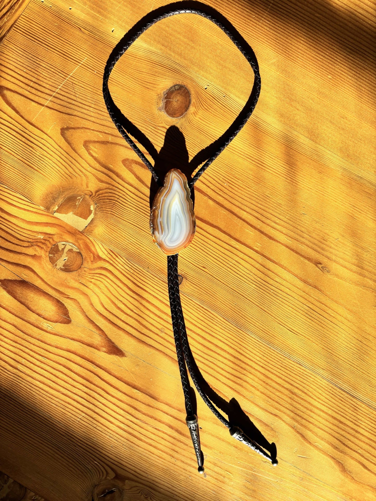 The Cimmaron Bolo Tie by My Store, featuring a polished natural banded agate stone centerpiece, rests on a sunlit wooden surface. Its braided leather cord and pewter decorative tips cast distinct shadows on the wood grain.