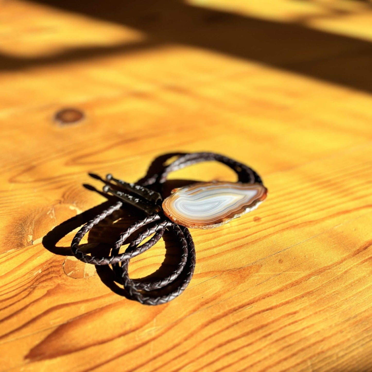 The Cimmaron Bolo Tie from My Store, featuring a polished banded agate centerpiece and dark braided leather cords, rests on a sunlit wooden table, casting soft shadows.
