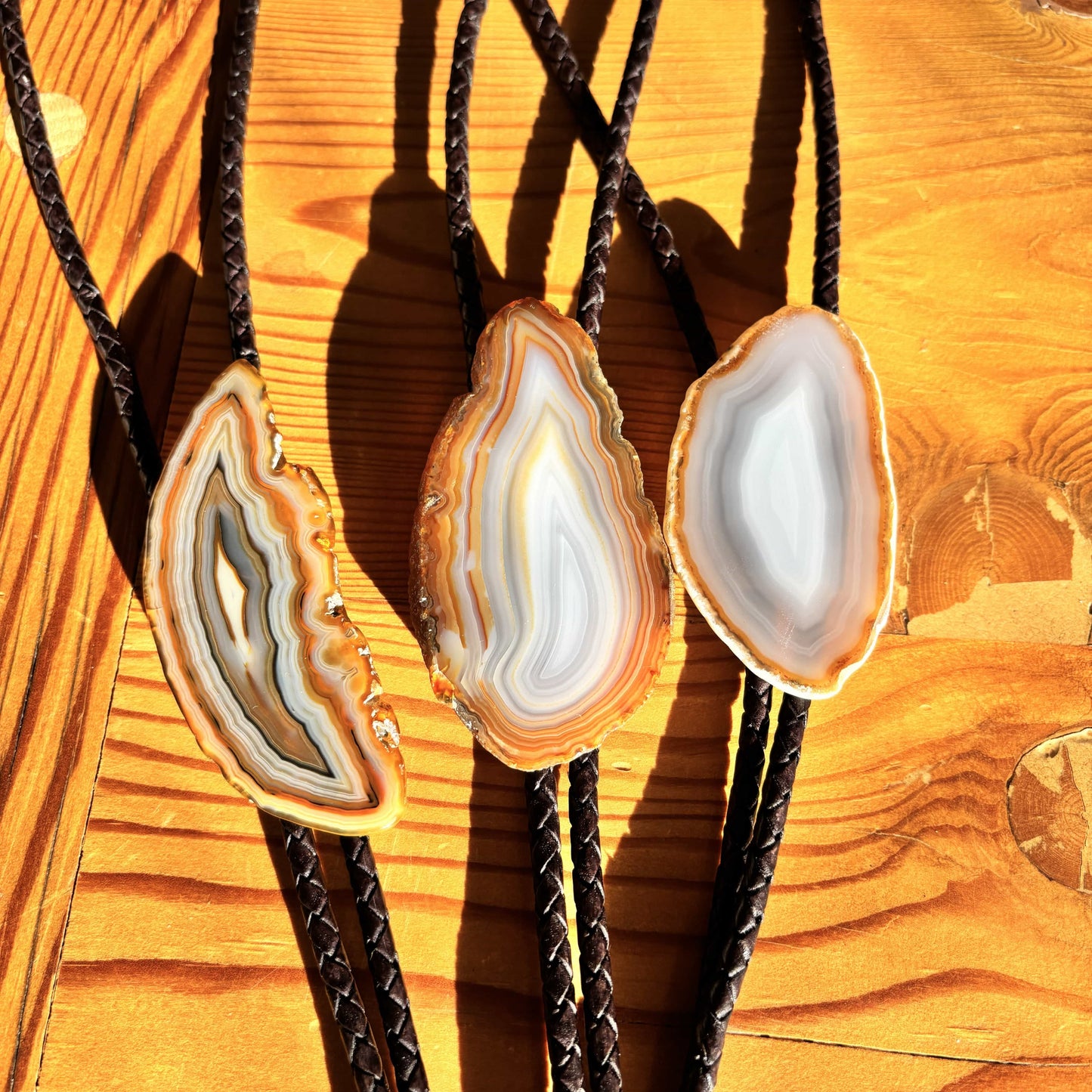Three Cimmaron Bolo Ties by My Store feature natural banded agate slides on a wooden surface, each with unique brown to white patterns. Braided leather cords with pewter tips cast sharp shadows in the sunlight.