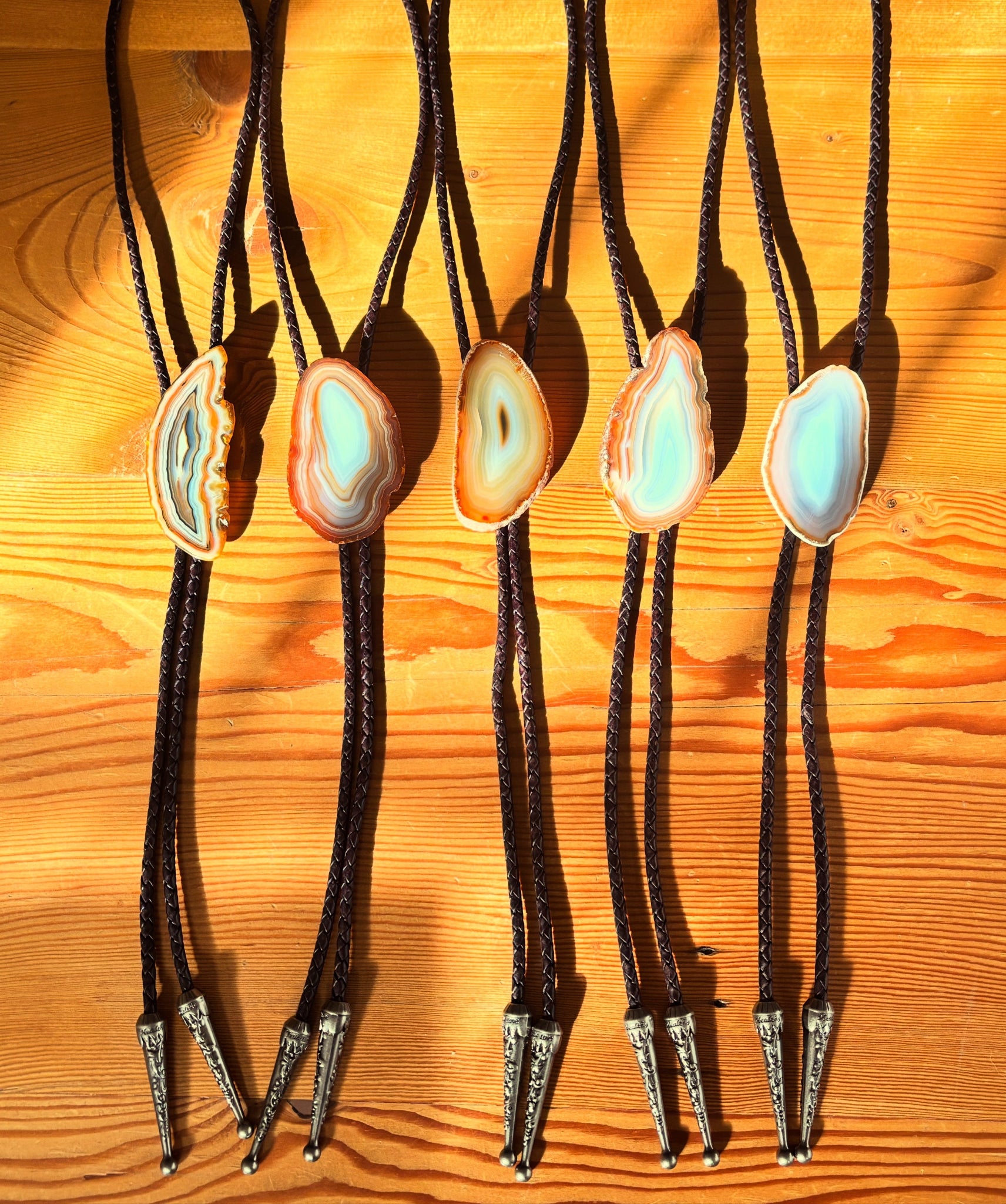 Five bolo ties with agate stone slides and leather cords are displayed on a wooden surface, casting shadows in bright sunlight.