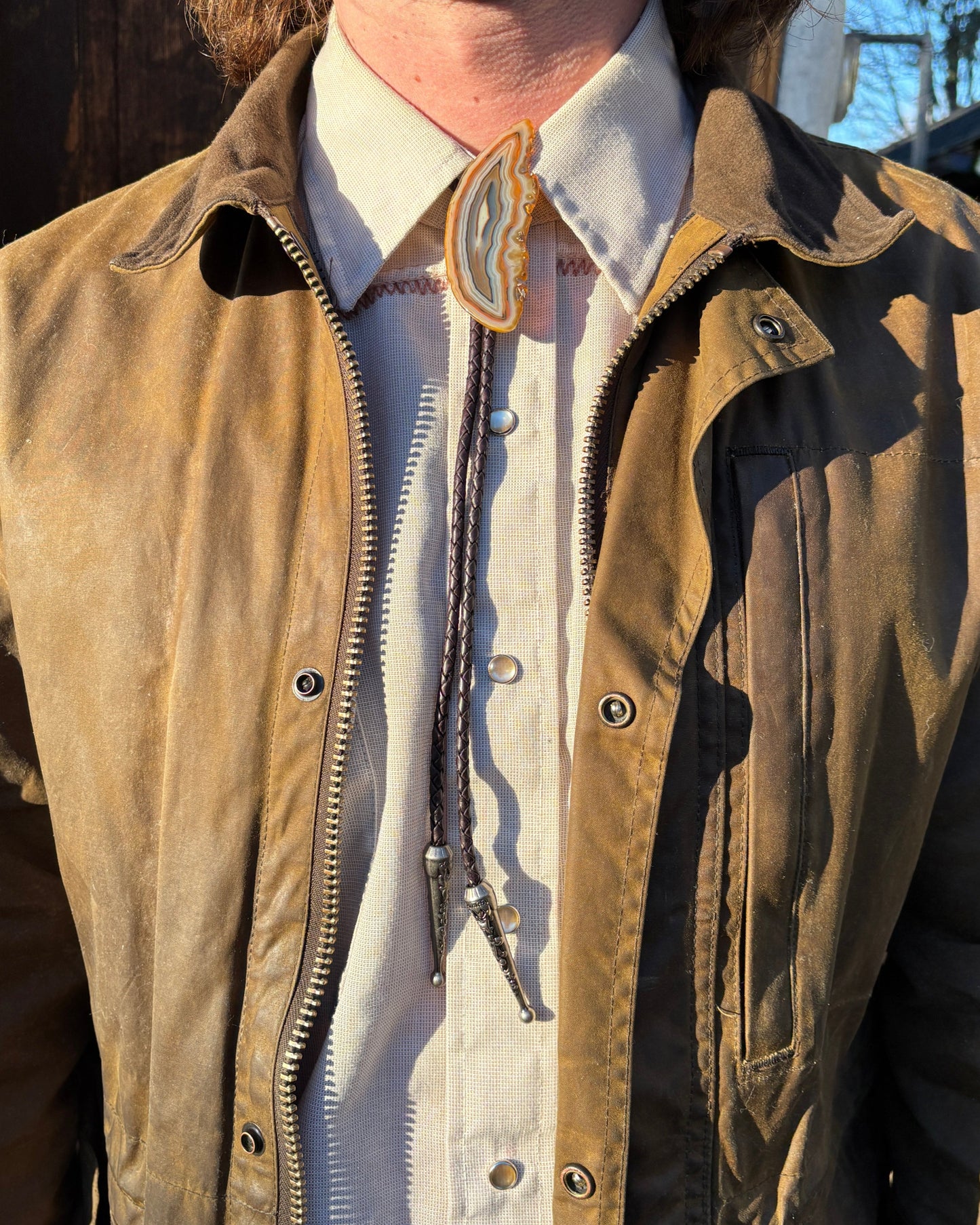 A person in a brown leather jacket over a beige button-up shirt is accessorized with the Cimmaron Bolo Tie from My Store, featuring natural banded agate and pewter tips. Sunlight highlights the clothings textures.