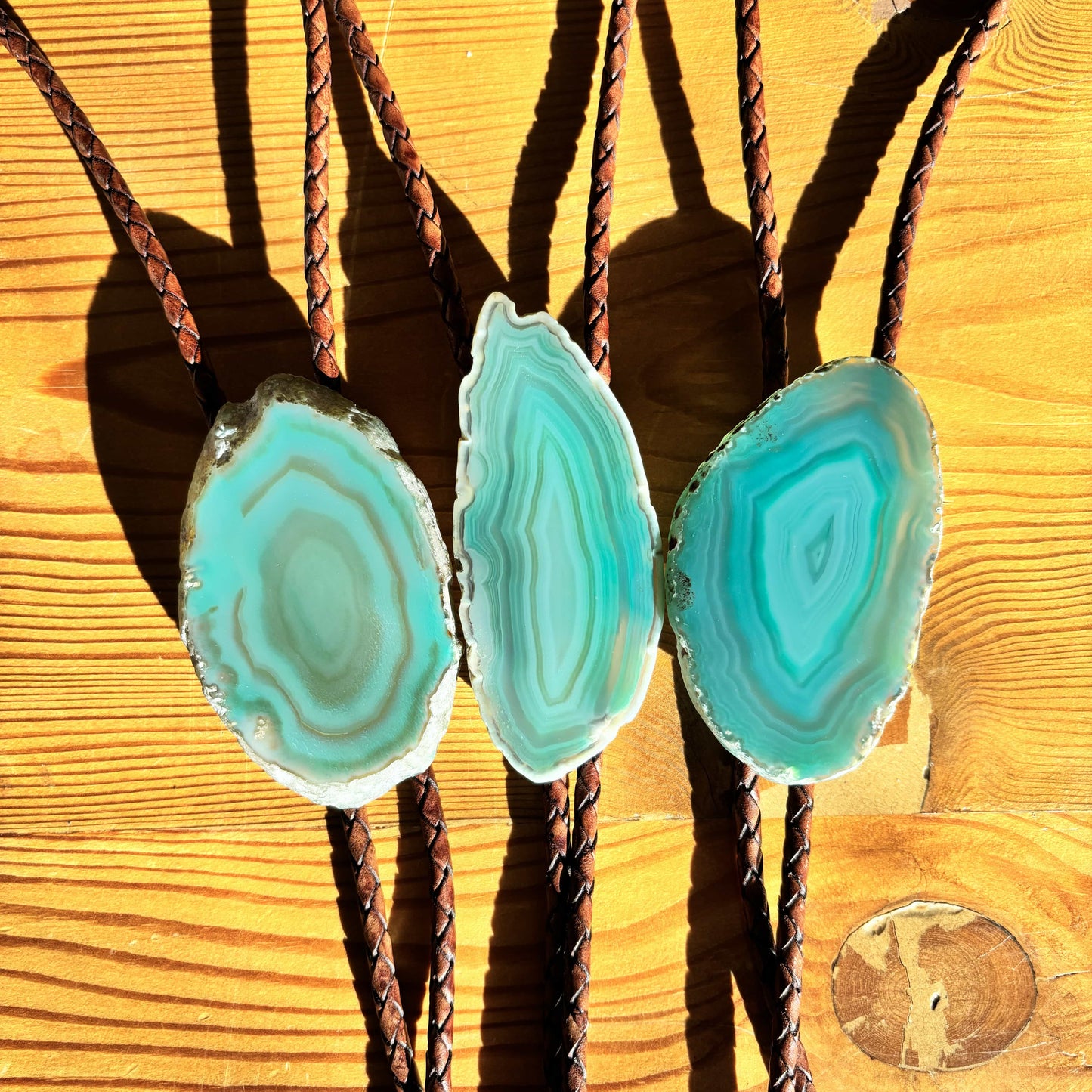 Three Sage Bolo Ties from My Store feature turquoise geode slices on a wooden surface. Each slice, attached to a handmade mahogany braided leather cord, casts soft shadows under warm lighting that beautifully highlights their intricate patterns and colors.