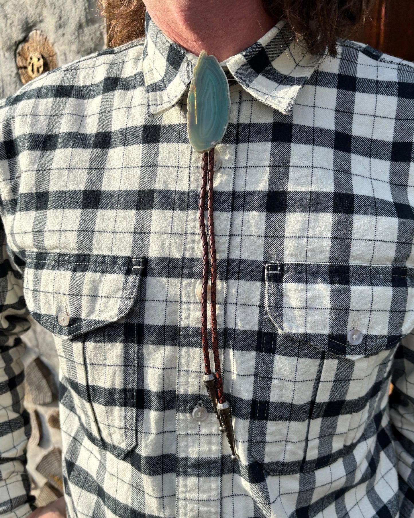 A person in a plaid shirt sports the handmade Sage Bolo Tie from My Store, featuring a large polished green banded agate stone clasp. The mahogany braided leather strings hang loosely, ending with metal tips.