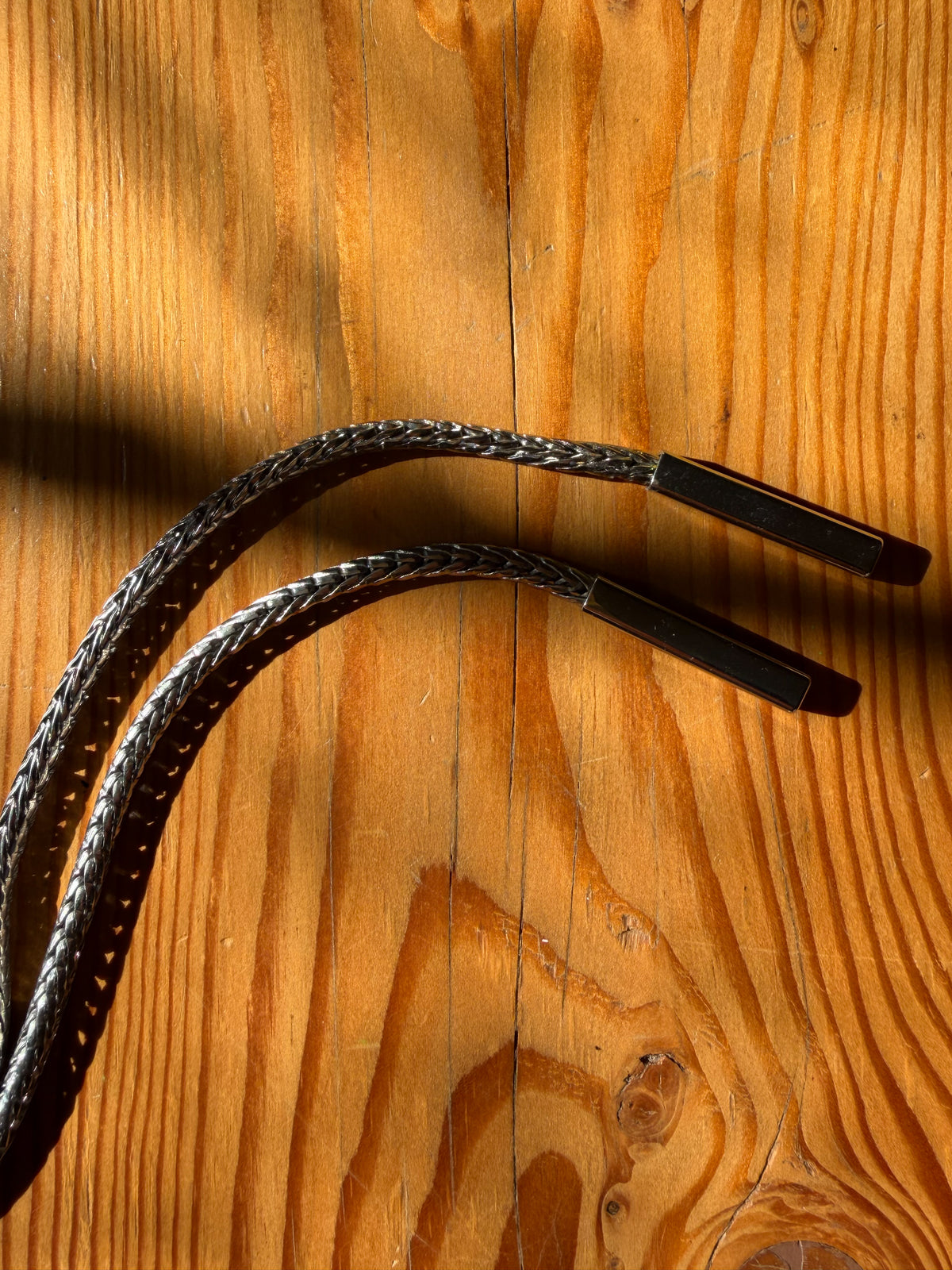 Two black braided audio cables with rectangular metal connectors, laid on a warm wooden surface with strong sunlight and shadows.