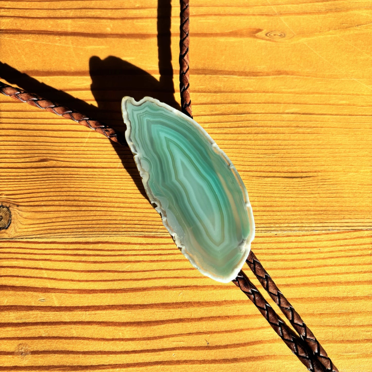 The Sage Bolo Tie from My Store showcases a green banded agate slice with white bands, set against a wooden surface to highlight the woods grain and knots. The agate is attached to a mahogany braided leather tie for a handmade touch.