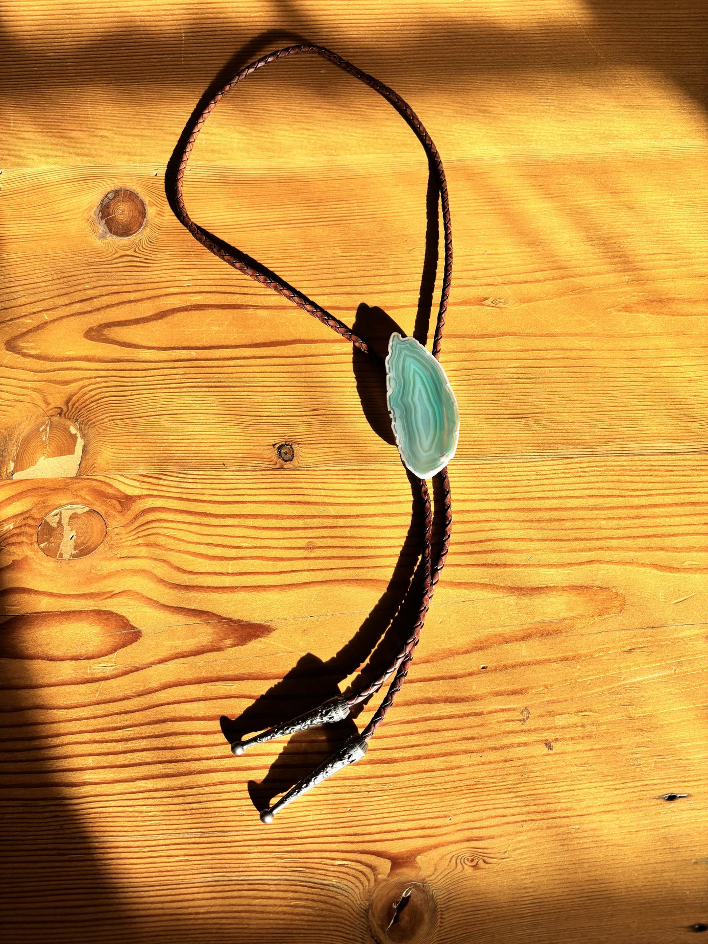 The Sage Bolo Tie by My Store, featuring a turquoise gemstone centerpiece on a mahogany braided leather cord with metal tips, is showcased on a wooden surface as warm sunlight casts shadows across the wood.