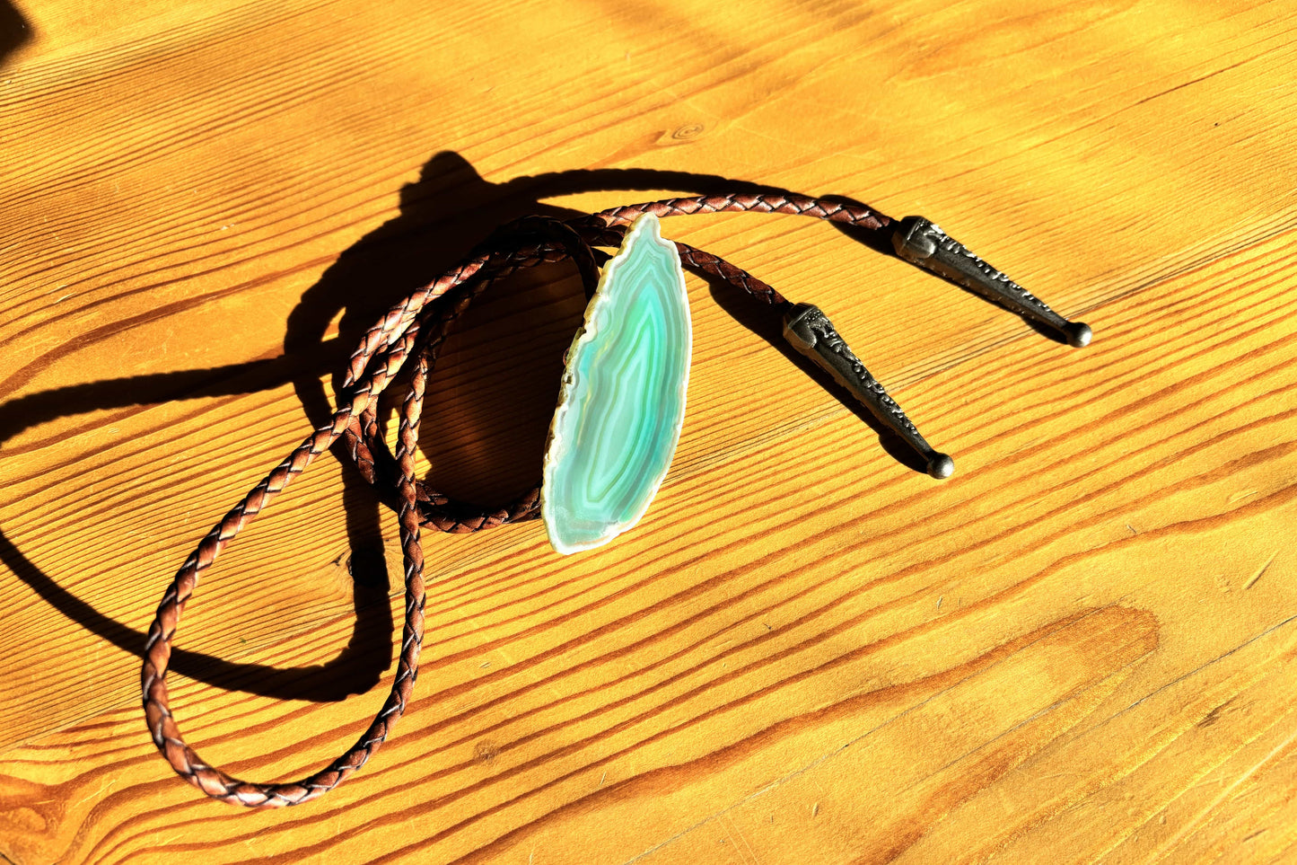 The Sage Bolo Tie from My Store, featuring a green banded agate stone centerpiece, rests on wood. Its mahogany braided leather cords with metal tips contrast the polished stones unique patterns as sunlight highlights its features.