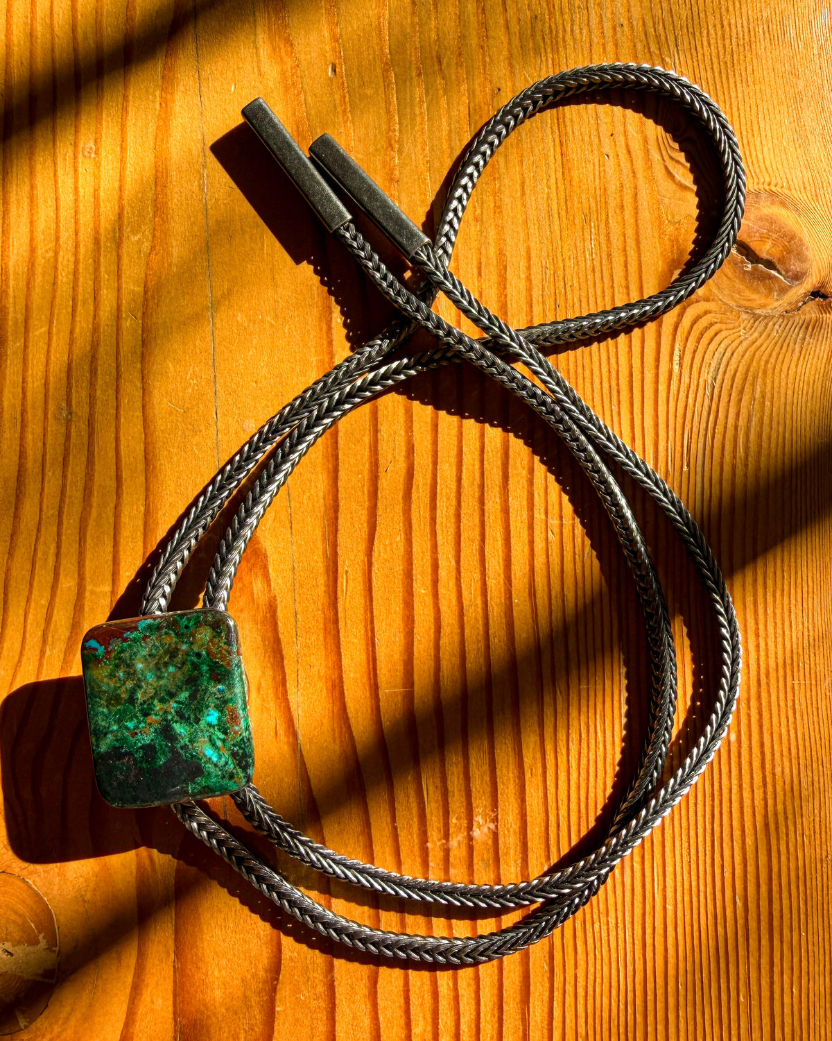 A braided metal bolo tie necklace with a square green and blue patinated stone clasp, displayed on a warm wooden surface with dramatic shadows.