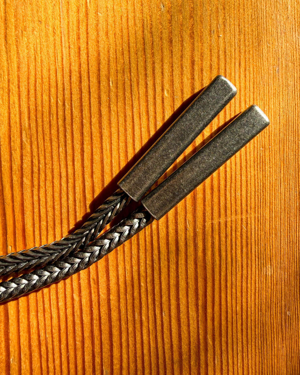A braided black and silver rope with two dark metal toggle sliders, laid diagonally on a warm orange-brown wood grain background.