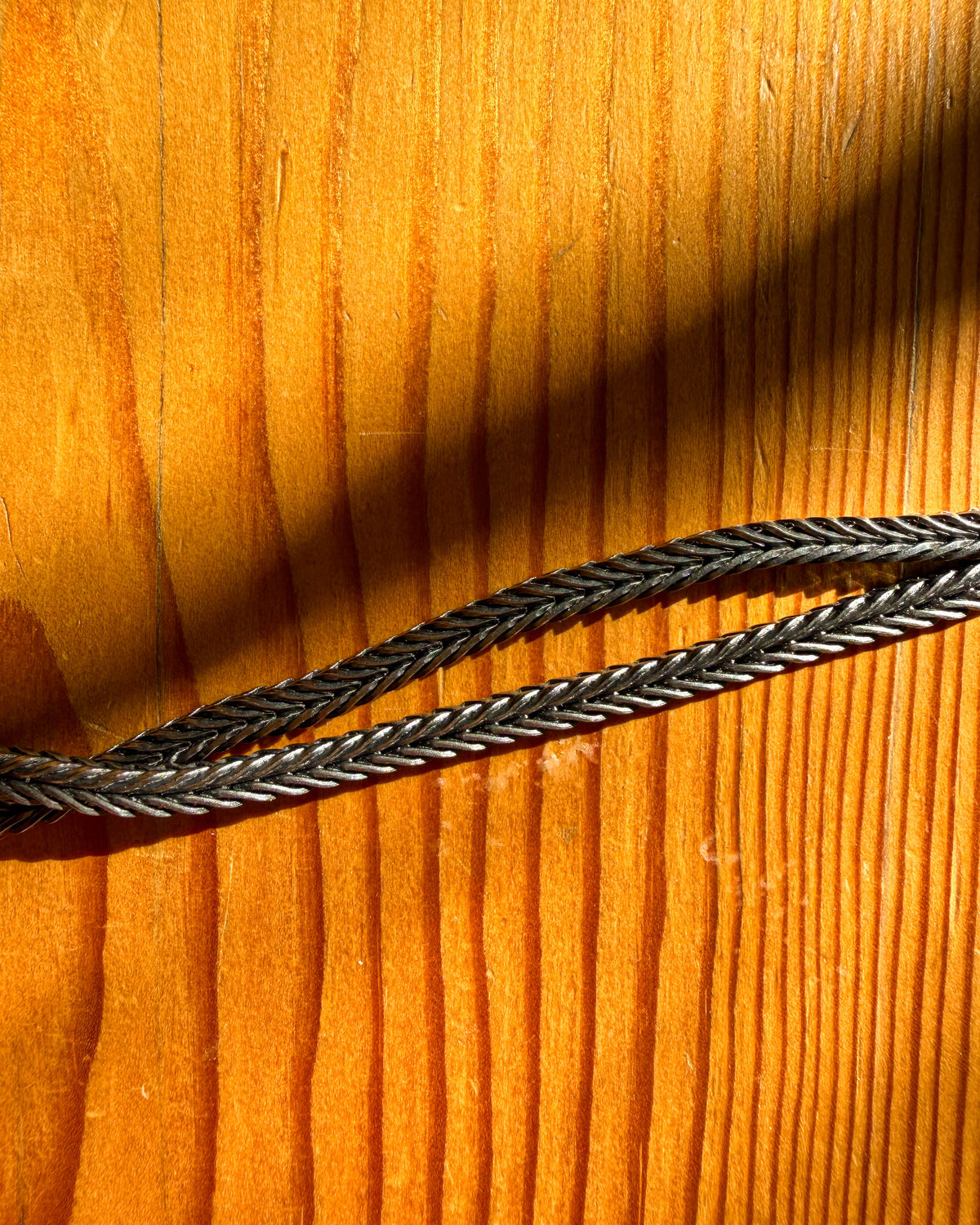 A close-up of a dark gray braided or woven strap laid diagonally across a warm orange-brown wood surface with strong sunlight and shadows.
