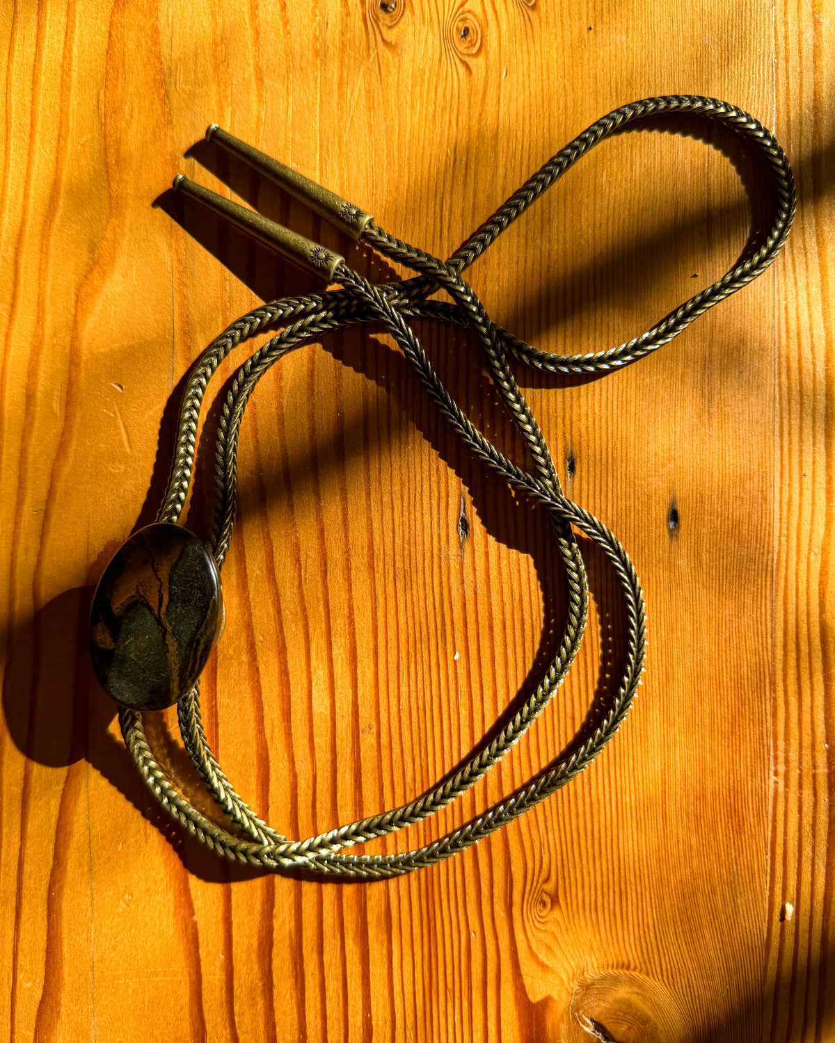 A metal brass chain necklace bolo tie with a polished oval stone pendant, laid out on a warm wooden surface with strong sunlight and shadows.