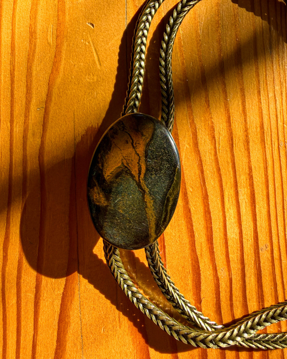 A close-up of a bolo-style necklace with a polished oval stone pendant and brass metal chain, laid on a warm wood surface.