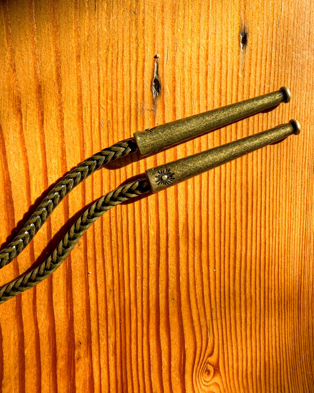 A close-up of a brass metal chain with brass metal tips and a decorative sun emblem, laid diagonally on a warm wood grain background.