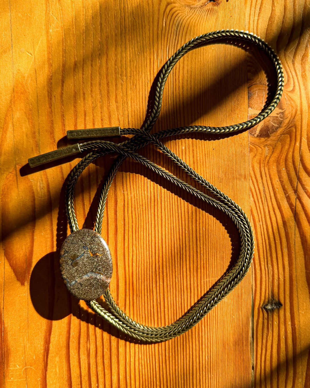 A braided metal bolo tie necklace with a round textured stone pendant, laid out on a warm wooden surface with strong sunlight and shadows.