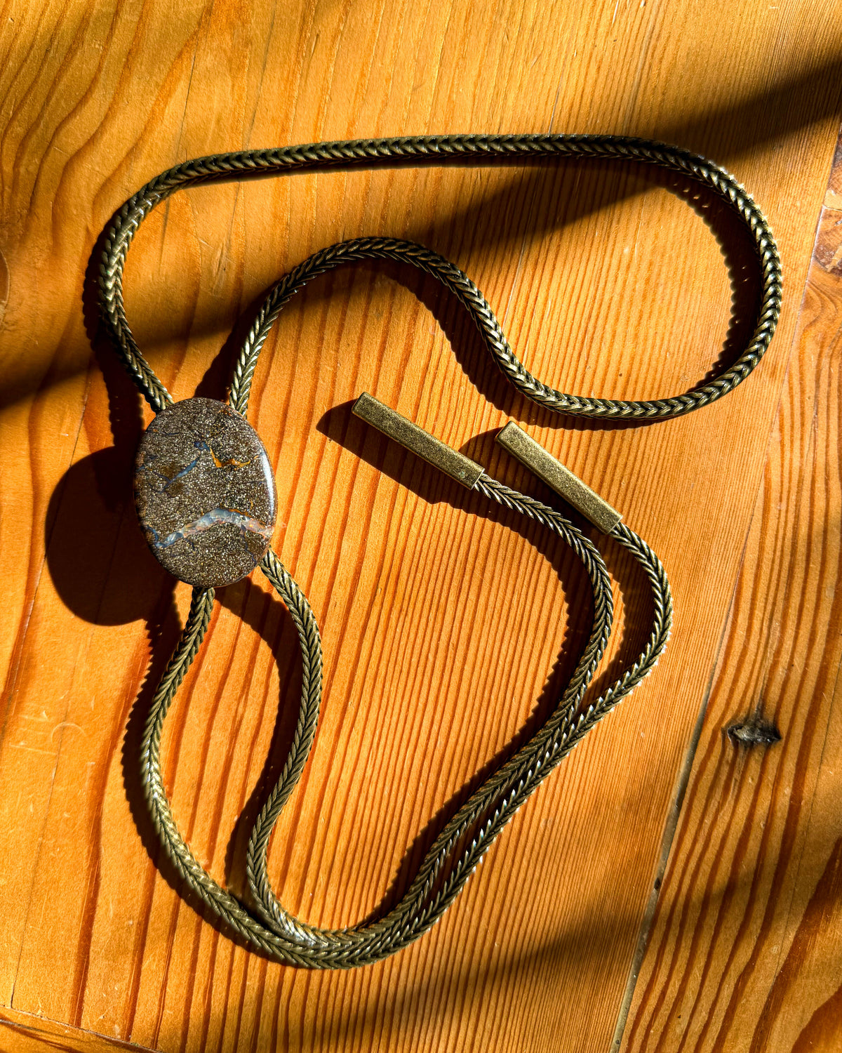 A vintage-style bronze-toned necklace with a textured oval pendant, laid out on a warm wooden surface with strong natural shadows.