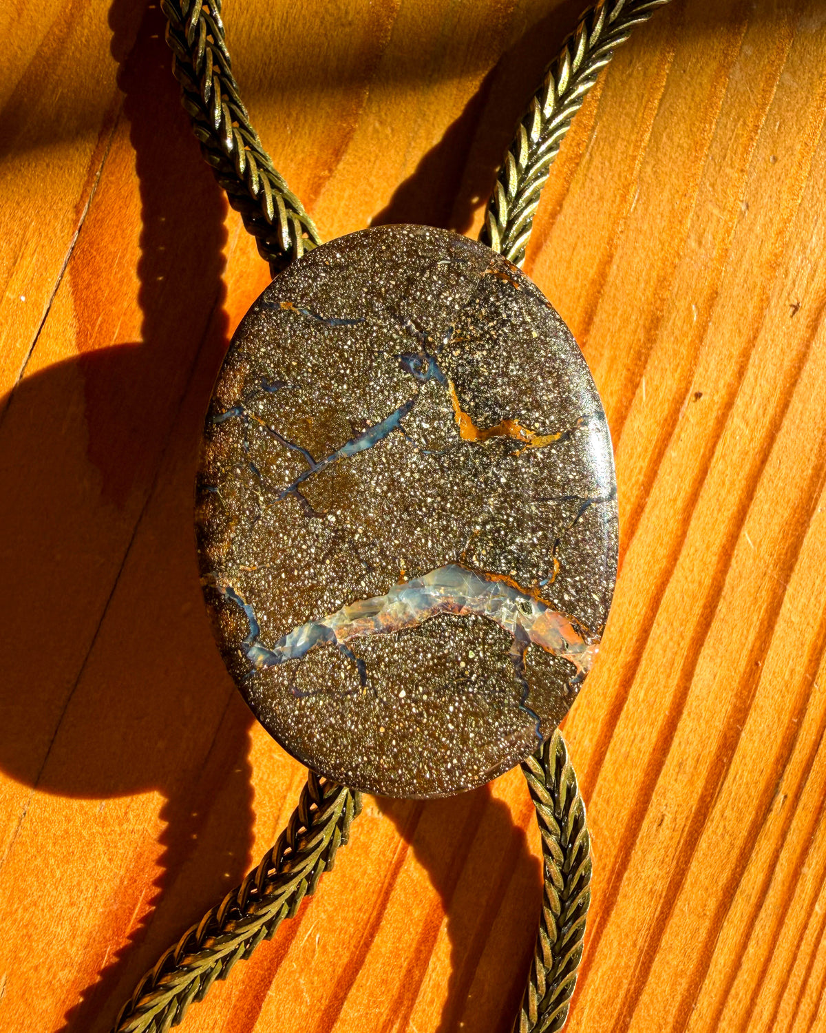 A close-up of a natural stone bolo tie pendant with brass braided metal cord, resting on a wooden surface under direct sunlight.
