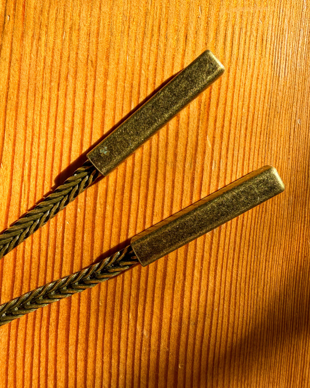 Close-up of braided metallic cords with rectangular metal end caps, placed diagonally on a warm orange-brown wood-grain surface.