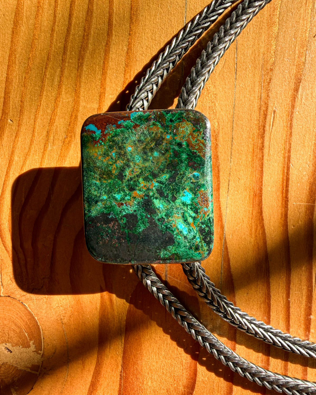 A close-up of a braided silver-toned bolo tie necklace with a rectangular green and turquoise stone pendant, laid on a warm wood surface.
