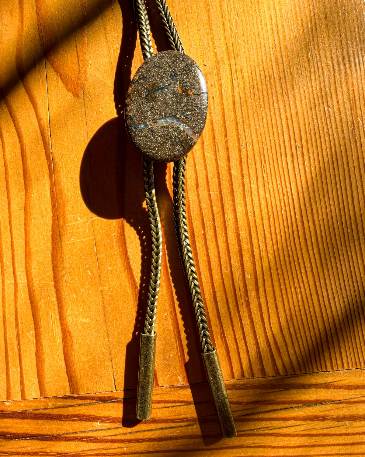 A natural stone bolo tie with braided cord and metal tips, displayed on a warm wooden surface with dramatic sunlight and shadows.