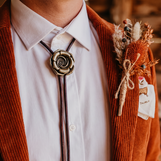 A close-up of a person wearing a dark brown leather bolo tie with a bronze rose-shaped metal clasp, styled with a white collared shirt and an rust-orange corduroy blazer. A rustic dried flower boutonniere is pinned to the left lapel.