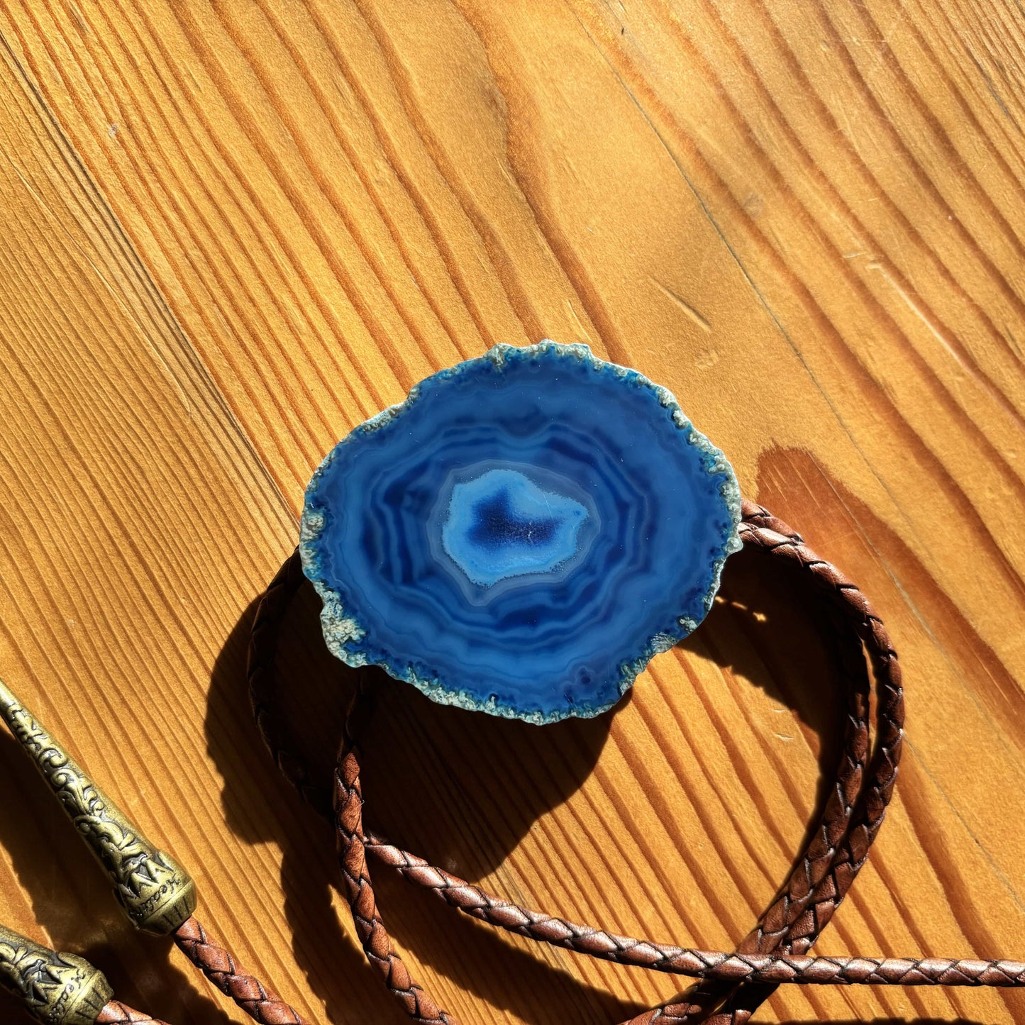 A vibrant Cobalt Bolo Tie from My Store, featuring a blue banded agate slice with concentric layers, is elegantly framed by decorative metal and braided leather. On a sunlit wooden surface, it casts distinct shadows that enhance its handmade charm.