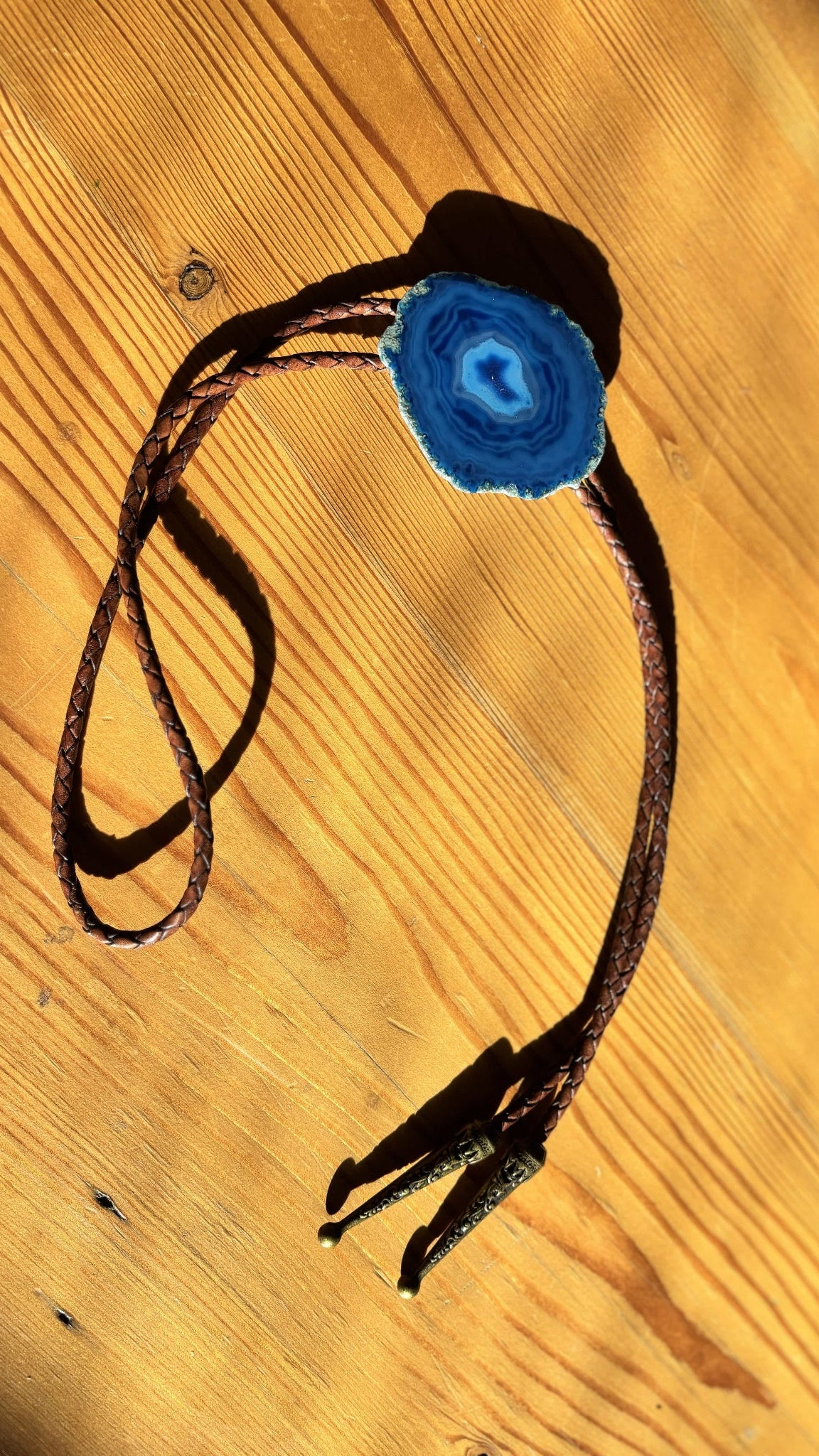 The Cobalt Bolo Tie by My Store, featuring braided leather cords and a blue banded agate stone centerpiece, rests on a wooden surface, casting soft shadows.