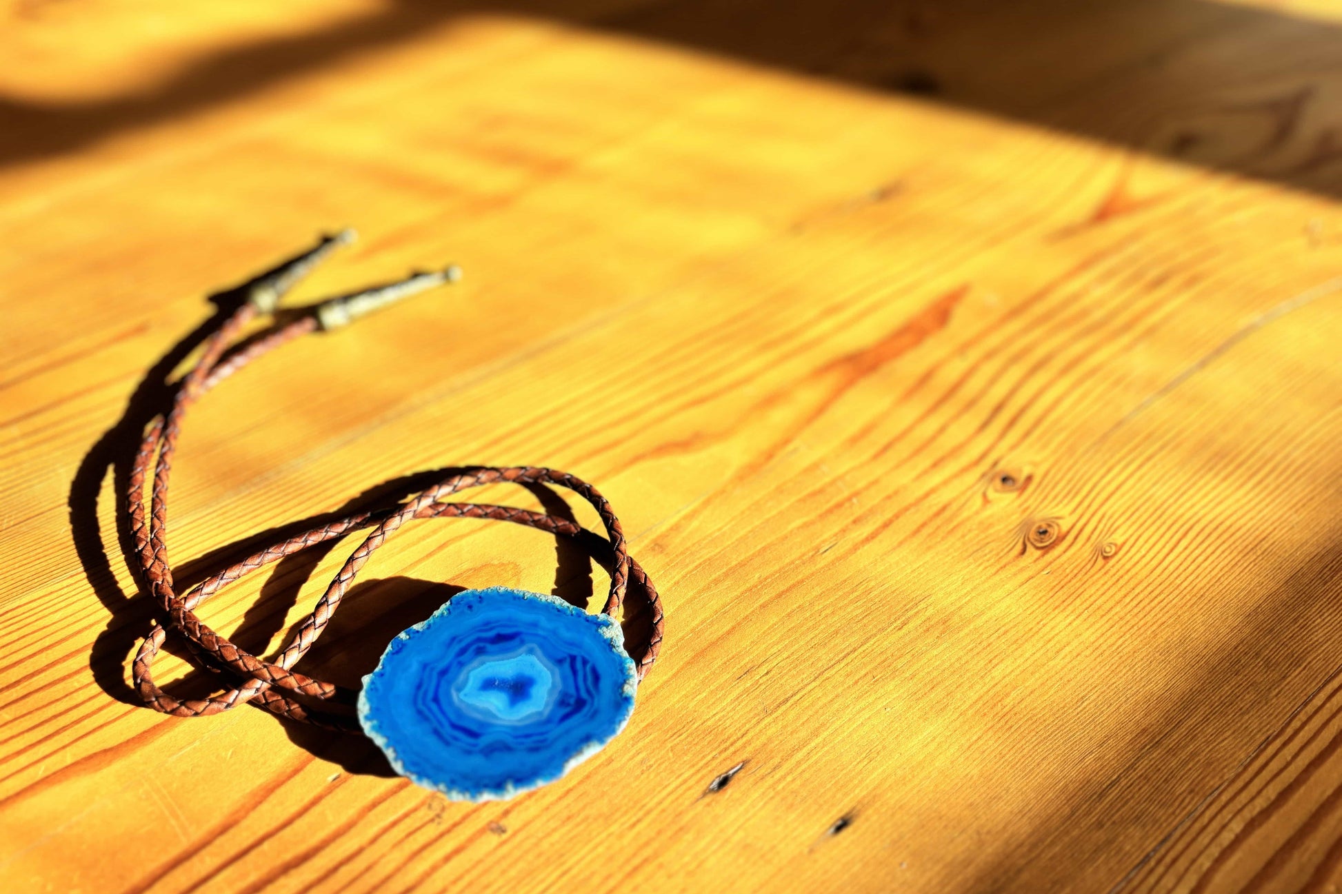 The Cobalt Bolo Tie by My Store features a blue banded agate slice pendant on a braided leather cord, elegantly displayed on warmly lit wood, with shadows accentuating the grains texture.