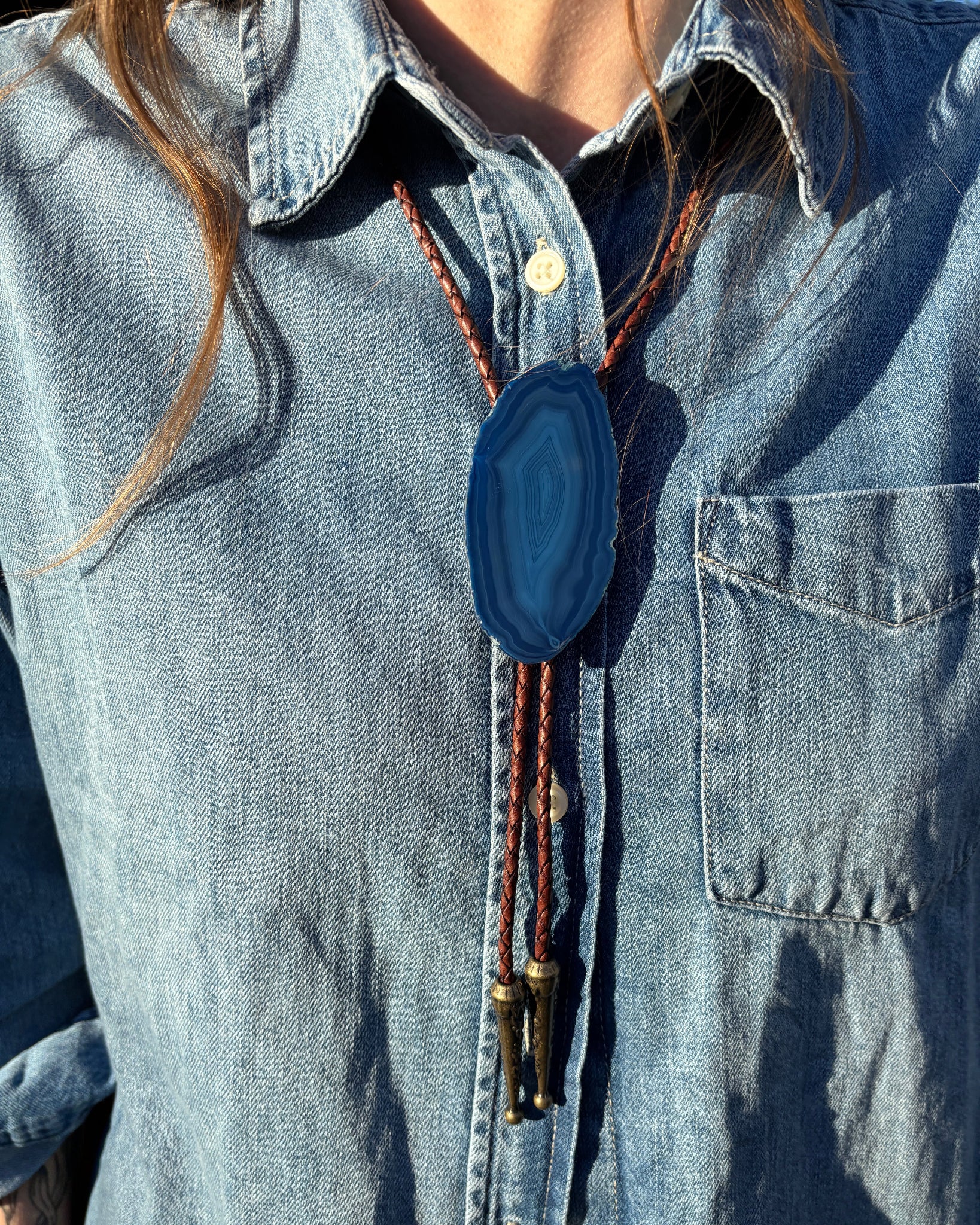 A person wears a denim shirt with the Cobalt Bolo Tie by My Store, featuring a striking blue banded agate pendant, intricately braided leather cords, and polished metal tips as sunlight dances on the shirt, casting playful shadows.