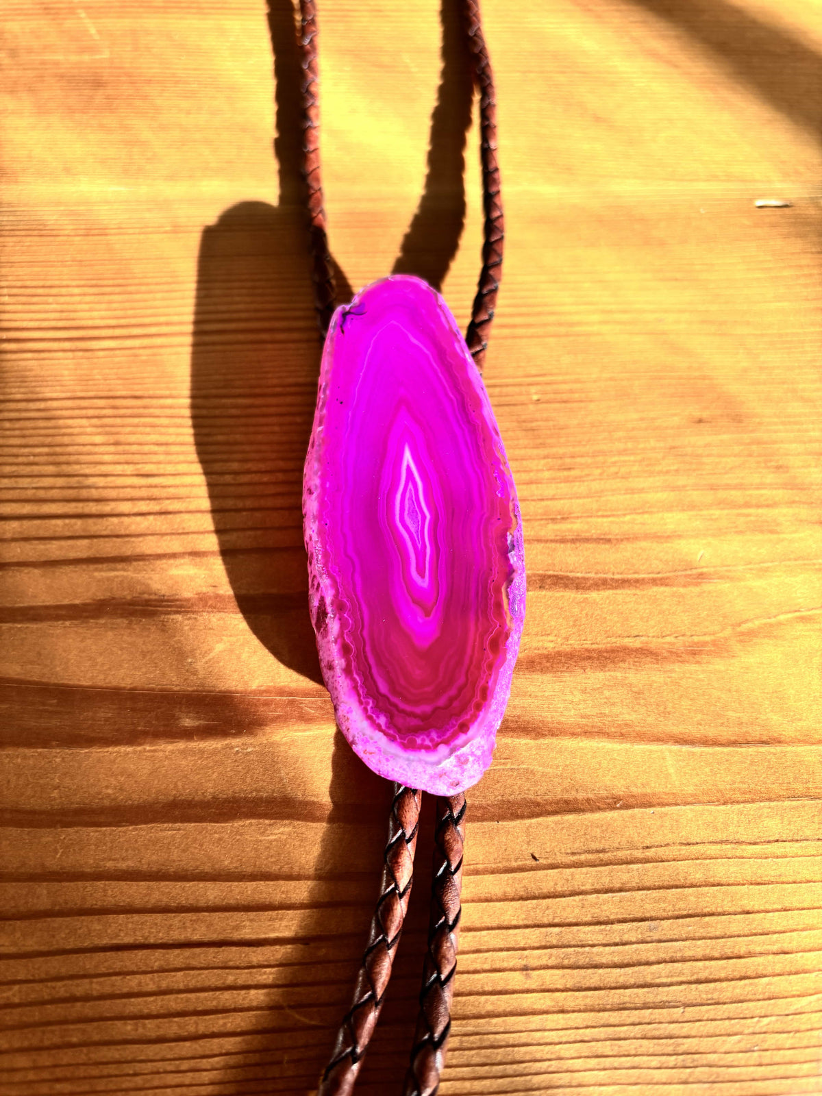 A vibrant pink geode pendant is set on braided leather cords, placed on a wooden surface. Sunlight casts shadows, highlighting the geodes concentric bands and rugged edges.