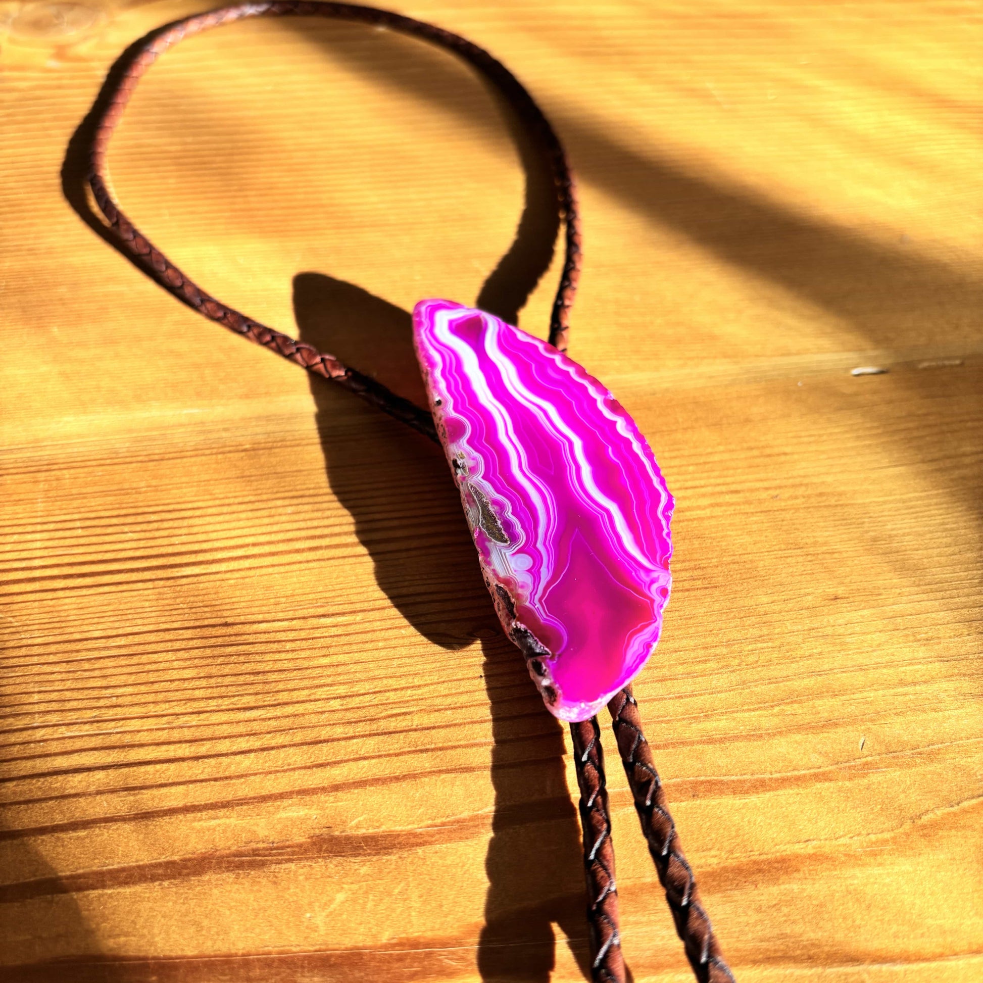 The Dolly Bolo Tie by My Store features a pink-banded agate slice pendant with white stripes on a mahogany braided leather cord, resting on a wooden surface where sunlight enhances the wood grains depth and highlights the stones vibrant color.