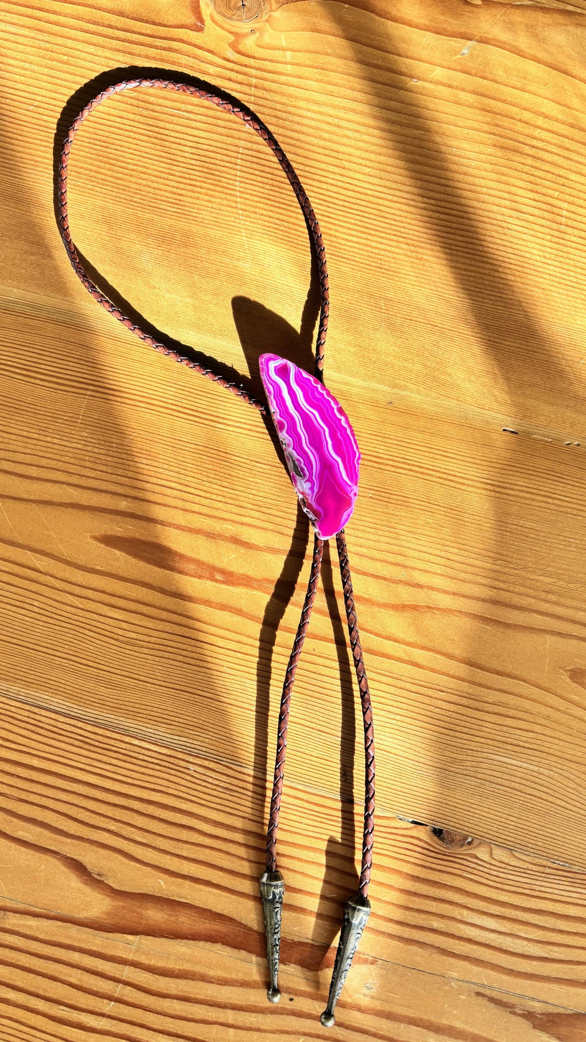 The Dolly Bolo Tie by My Store, featuring a pink banded agate stone and mahogany braided leather cords with silver metal tips, rests on wood. Sunlight casts elegant shadows over the scene.