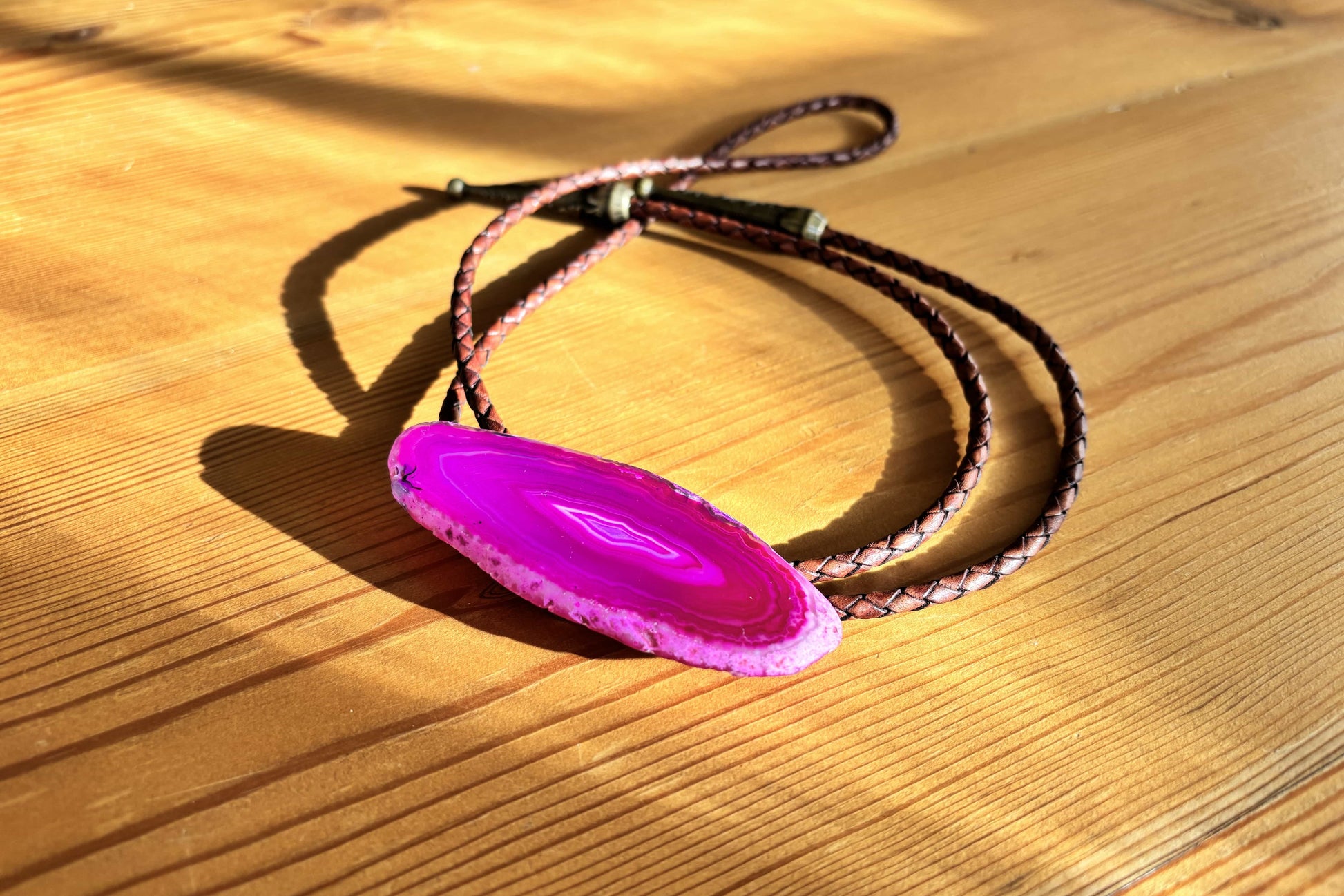 The Dolly Bolo Tie by My Store features a vibrant pink banded agate pendant on a mahogany braided leather cord, resting on wood. Sunlight casts a shadow highlighting the geodes concentric patterns.