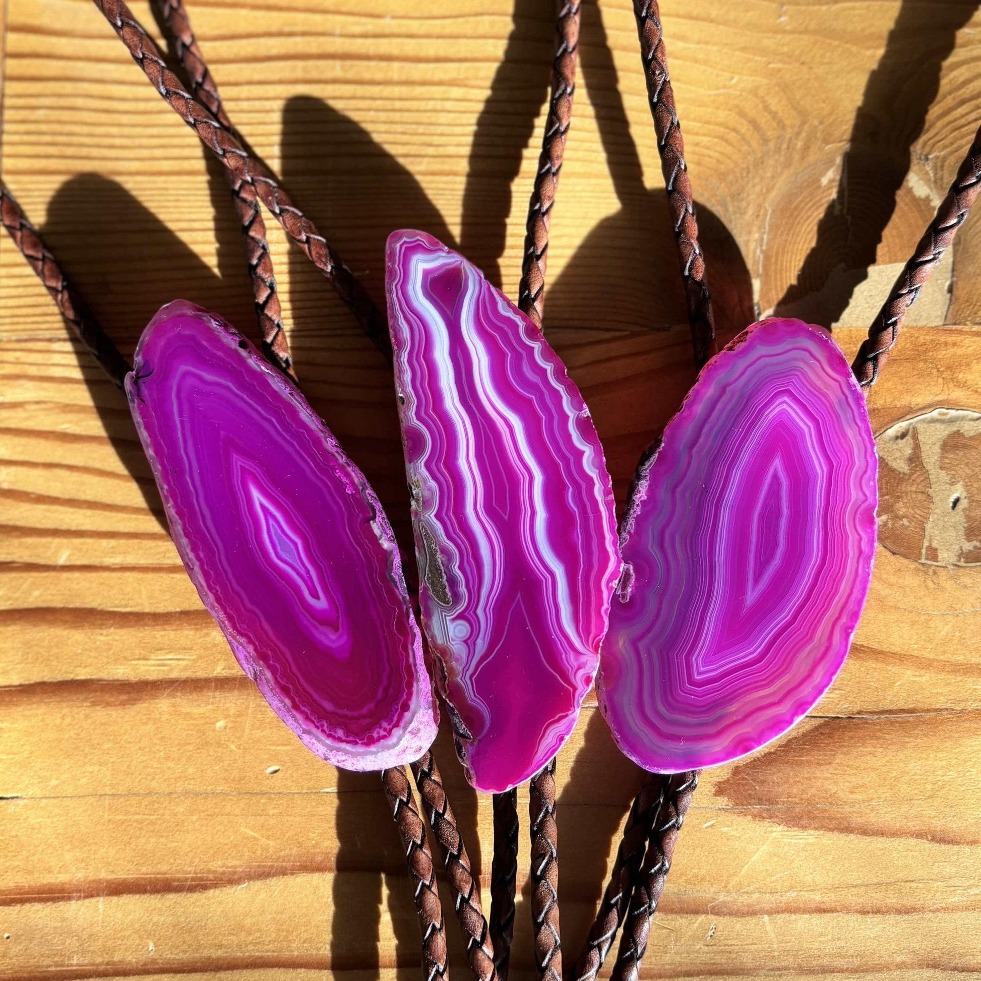 The Dolly Bolo Tie from My Store features three vivid pink banded agate slices with intricate patterns on a wooden surface, set against mahogany braided leather cords. The woods rustic texture beautifully contrasts the stones, creating a stunning handmade-inspired display.