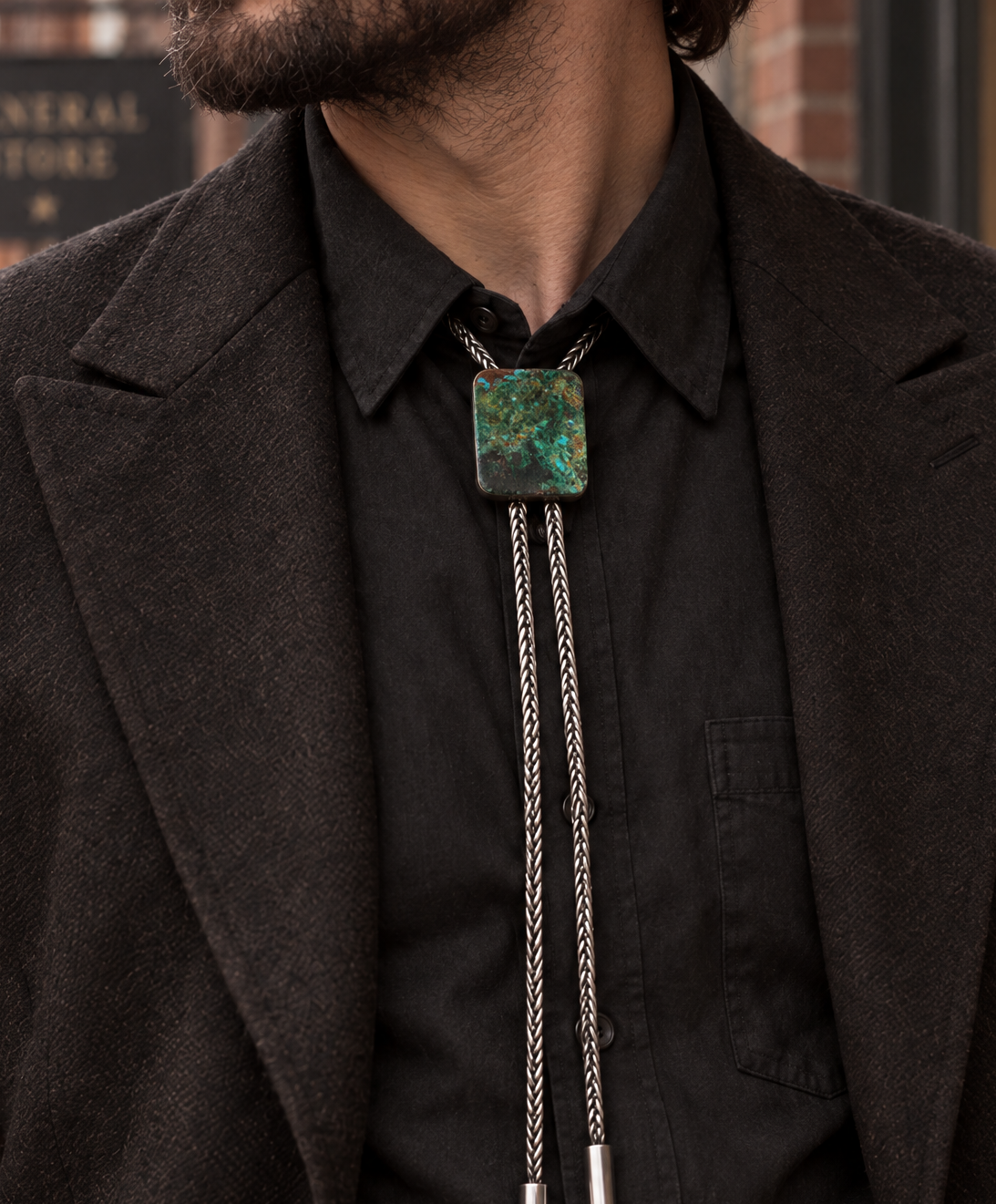 A close-up of a man wearing a silver-toned bolo tie with a square green and blue marbled stone pendant, styled over a black collared shirt and dark brown blazer.