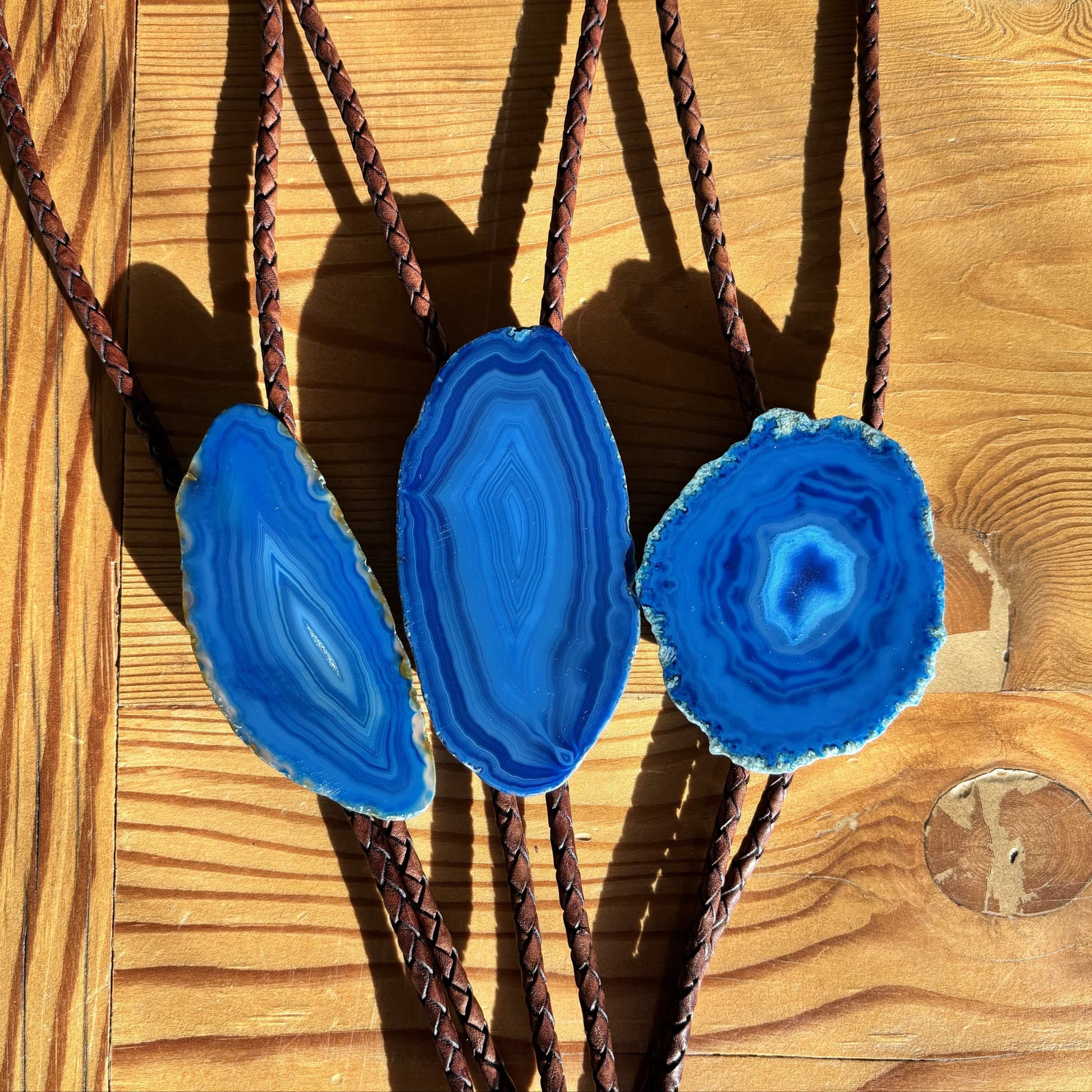 On a wooden surface, three Cobalt Bolo Ties from My Store feature uniquely patterned blue banded agate stones. Their braided leather cords enhance the agates vibrant hues, creating a striking visual harmony.