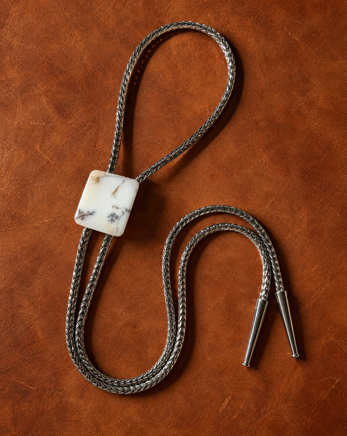 A braided silver-toned bolo tie necklace with a square white stone slide featuring black and gray marbling, displayed on a textured brown leather background.