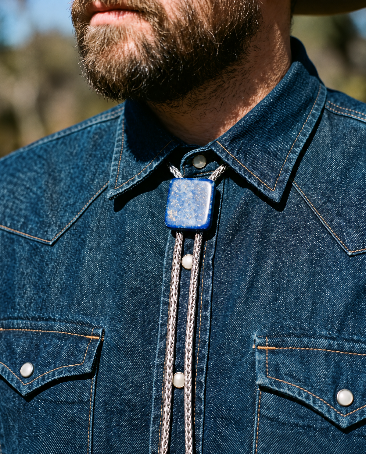 A close-up of a man wearing a dark blue denim western shirt and a blue square bolo tie with braided silver cords.