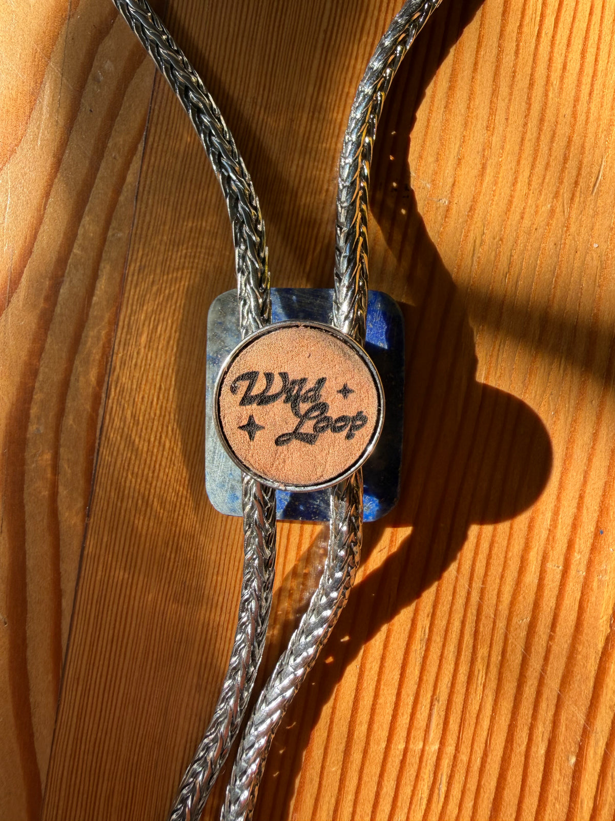 A close-up of a bolo tie necklace with braided silver-toned cords, a square blue stone pendant, and a round tan leather center disc engraved with the words 'Wild Loop' in black script, resting on a wooden surface.