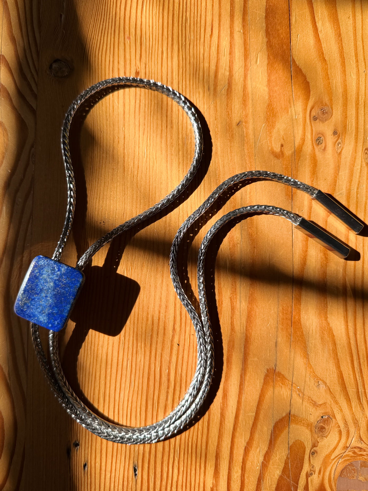 A braided silver-toned necklace with a square blue lapis lazuli pendant, laid out on a warm wooden surface with strong sunlight and shadows.