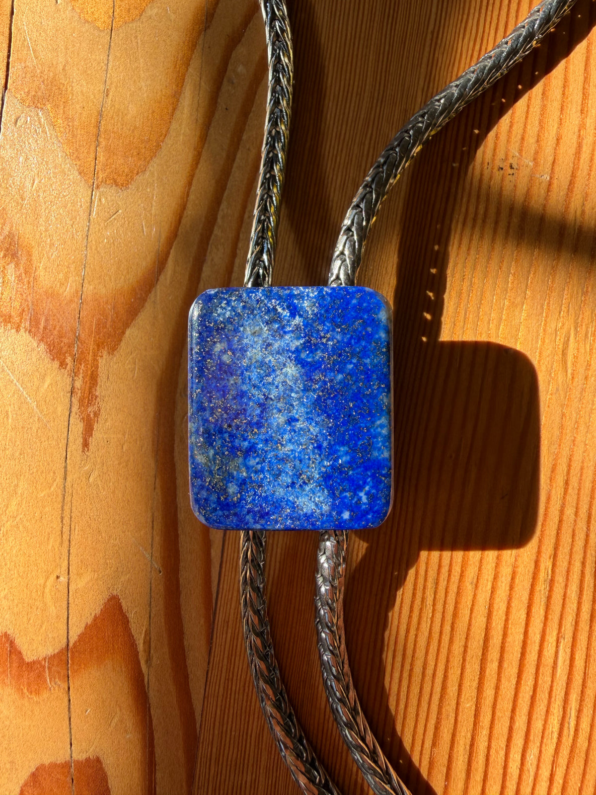 A square blue lapis lazuli stone pendant necklace with a braided silver-toned cord, displayed on a warm wooden surface.