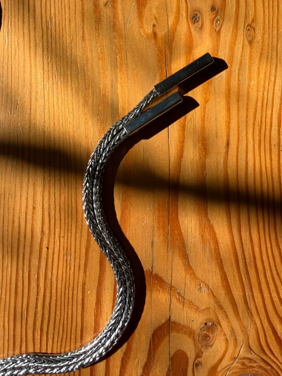 A close-up of a braided silver cable with black rectangular connectors, laid on a warm wood surface with strong sunlight and shadows.