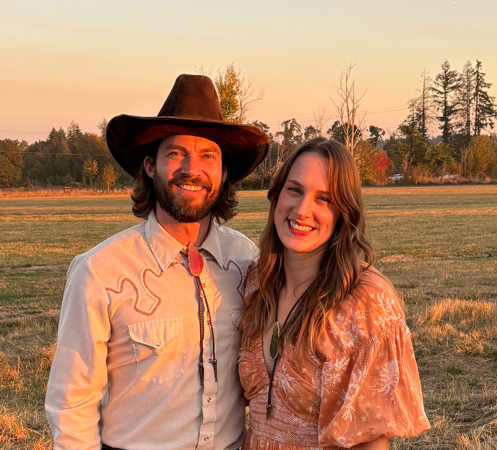A man and woman stand in a field at sunset, smiling. The man wears a cowboy hat and Western-style shirt. The woman is in a patterned dress. Tall trees and a warm sky form the background.