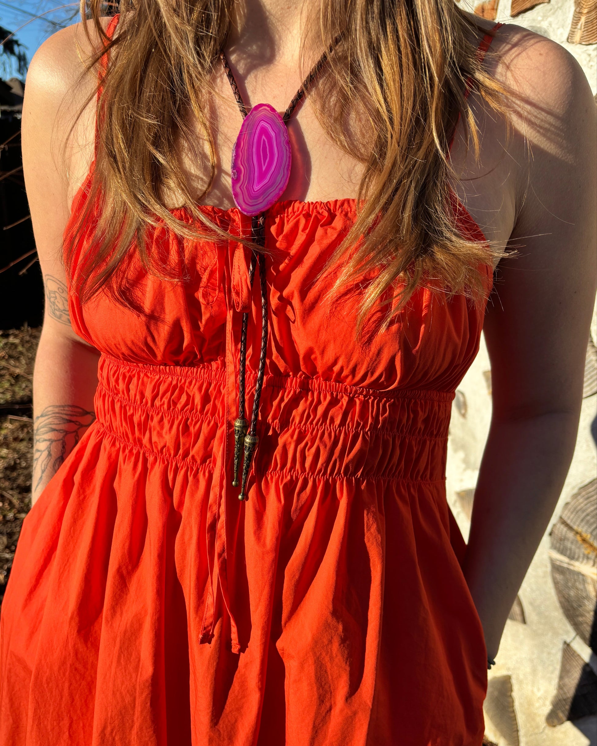 A person in a vibrant orange dress with a gathered waist wears My Stores Dolly Bolo Tie and a striking purple geode necklace. Their long hair flows elegantly against a sunlit backdrop of trees and leaves.